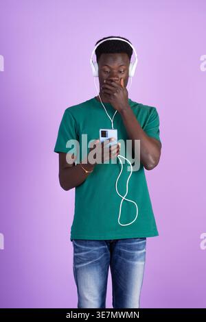 Shocked Black man in green shirt wearing headphones covering mouth with hand looking at smartphone standing against purple background in studio Stock Photo