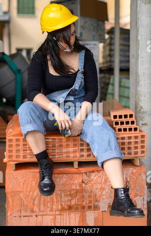 Serious Hispanic woman wearing denim overalls and yellow hardhat looking down with hair covering half face sitting on terracotta bricks at construc... Stock Photo