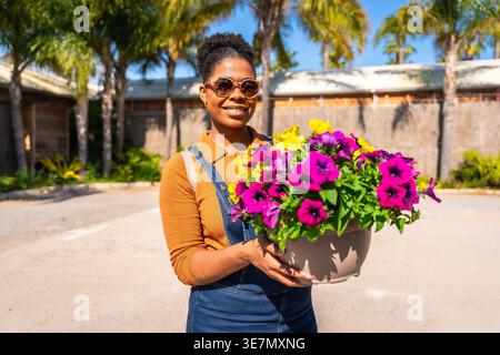 Hispanic woman in denim overalls and sunglasses holding potted flowers while standing outdoors on sunny day Stock Photo