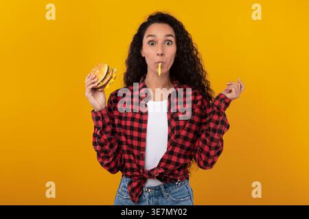 Middle Eastern woman with curly hair holding burger standing with fries in mouth against yellow background in studio Stock Photo