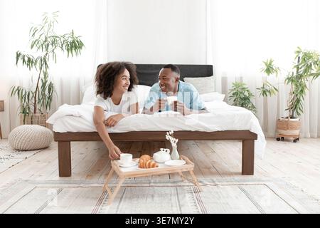 Smiling Black couple looking at each other having breakfast holding cup of coffee while lying on bed in bedroom at home Stock Photo