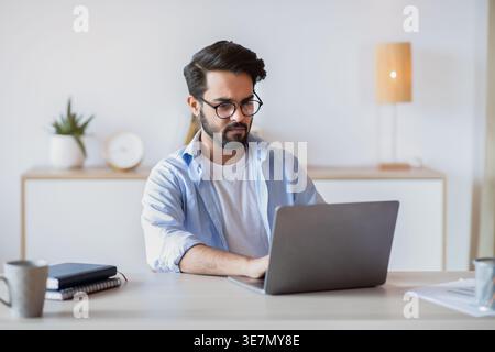 Telecommuting Concept. Focused Middle Eastern Man Using Laptop , Sitting At Desk In Home Office, Handsome Arab Freelancer In Eyeglasses Typing On Keyb Stock Photo
