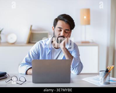 Middle Eastern man with closed eyes leaning face on hand sitting at table at home office Stock Photo