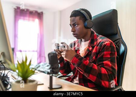 Black male gamer wearing headphones using joystick playing video games while sitting at table with microphone at home Stock Photo