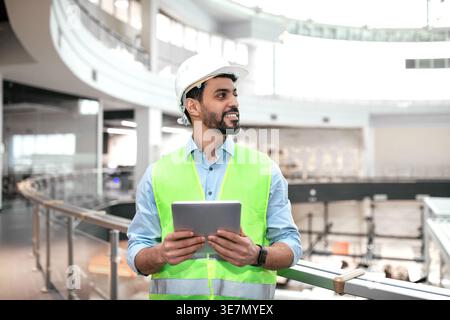 Middle Eastern male engineer wearing hard hat and vest looking away and holding tablet while standing beside terrace in office Stock Photo