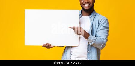 Black man pointing to blank whiteboard in his hands standing against yellow background in studio Stock Photo