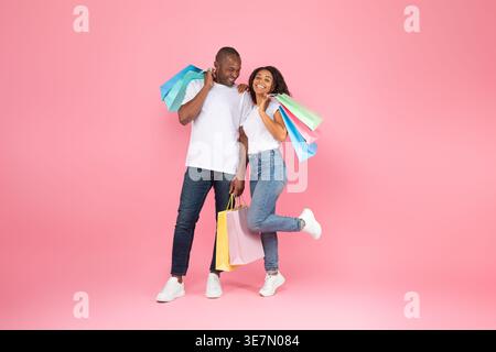 Smiling Black couple with colorful shopping bags standing against pink background in studio Stock Photo