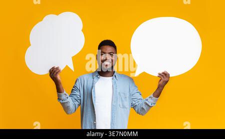 Happy Black guy holding two speech bubbles on yellow background at studio Stock Photo