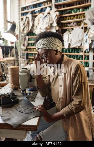 Black female tailor wearing eyeglasses touching forehead while sitting at table with leather in workshop Stock Photo
