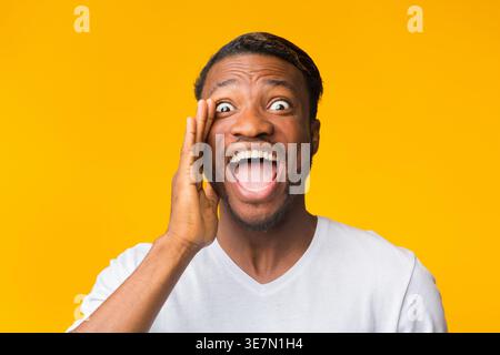 Black man in white shirt with mouth wide open touching face standing against yellow background in studio Stock Photo