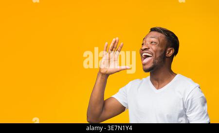Attention Concept. Black Man Shouting Holding Hand Near Mouth Standing Over Yellow Background In Studio. Panorama, Free Space Stock Photo