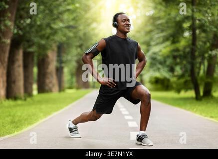 Black man in activewear wearing headphones doing stretching on road in forest during daytime Stock Photo