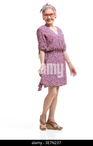 Smiling Chinese senior woman in floral dress with eyeglasses standing against white background in studio Stock Photo