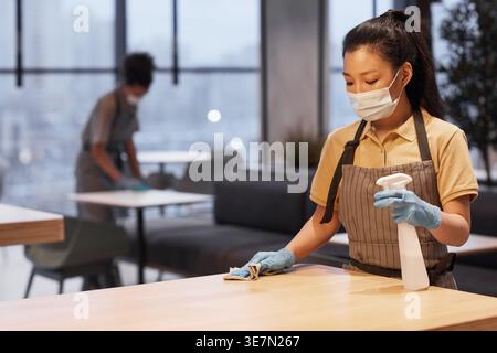 Asian female waitress wearing face mask and apron holding spray bottle and cleaning tables while standing in cafe during pandemic Stock Photo
