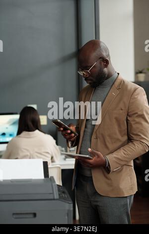 Black businessman using smartphone and digital tablet while standing in front of xerox machine in office Stock Photo