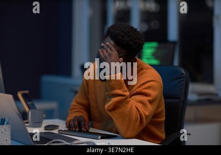 Tired Black male programmer touching forehead sitting at table with laptop working late at night in office Stock Photo