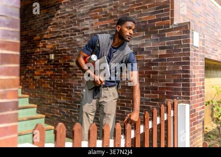 Black man holding water bottle and laptop leaving home on sunny day Stock Photo