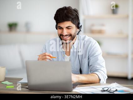 Smiling Arab man with beard wearing headset having video call through laptop while sitting at desk at home Stock Photo