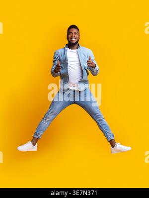 Cheerful Black man gesturing thumbs up while jumping against yellow background in studio Stock Photo