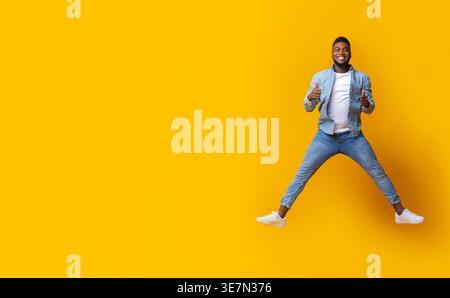 Cheerful Black man gesturing thumbs up jumping in the air against yellow background in studio Stock Photo