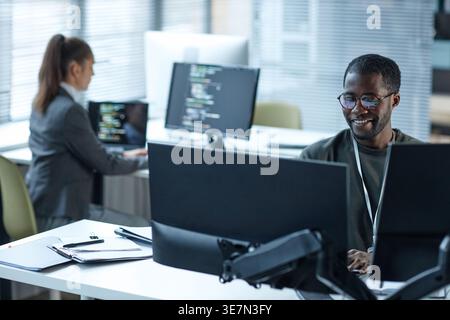 Smiling Black male programmer wearing eyeglasses working on computer sitting at table near caucasian female colleague in office Stock Photo