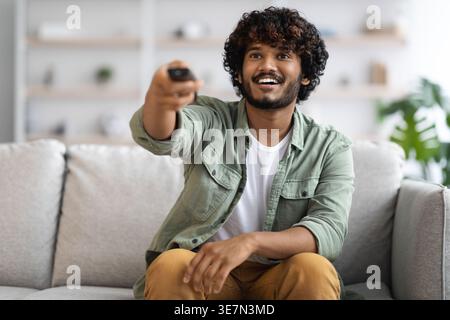 Asian man with curly hair using remote watching movie while sitting on sofa in living room at home Stock Photo