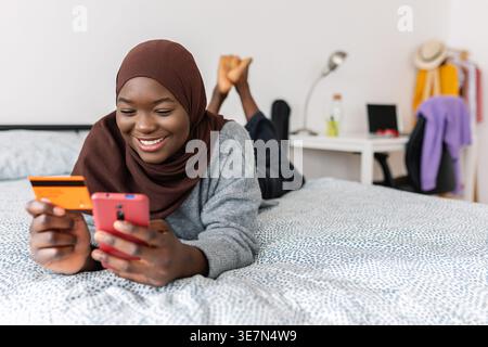 Black Muslim female teenager in brown hijab shopping online using credit card and smartphone while lying down on bed at home Stock Photo