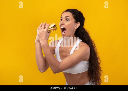 Middle Eastern woman with curly hair wearing sportswear holding and looking at burger while standing against yellow background in studio Stock Photo