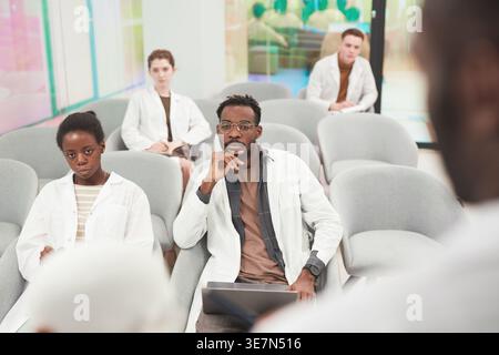 Diverse group of medical students in lab coats using laptop and taking note while listening to lecture during medical seminar in conference room Stock Photo