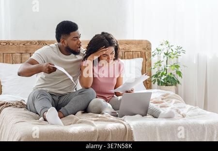 Black husband and wife checking documents suffering problems with managing budget while sitting together on bed with laptop at home Stock Photo