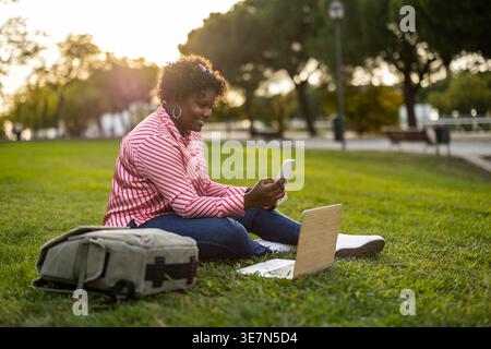 Curvy African American woman with curly hair using smartphone sitting on grass near backpack and laptop in park at sunset Stock Photo