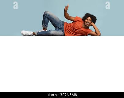 Smiling Black man with afro showing thumbs up lying on huge white board against blue background in studio Stock Photo