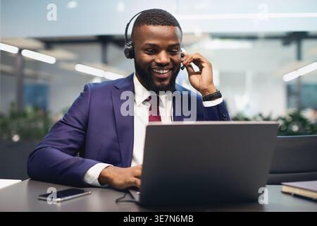 Smiling Black businessman in suit wearing headset having video conference on laptop sitting at table in office Stock Photo