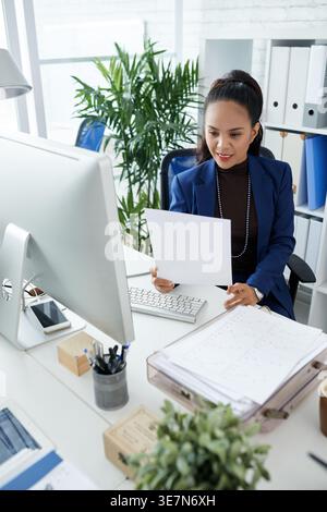 Smiling Filipino businesswoman holding documents while sitting at table with monitor in office Stock Photo