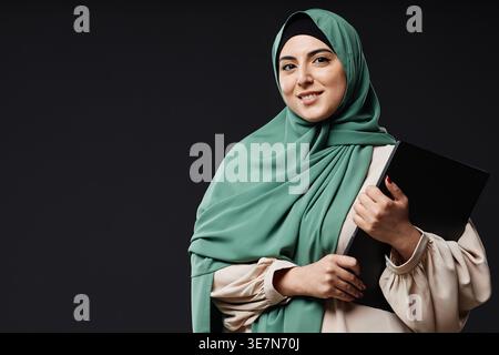 Smiling mixed-race woman wearing hijab carrying laptop while standing against black wall in studio Stock Photo