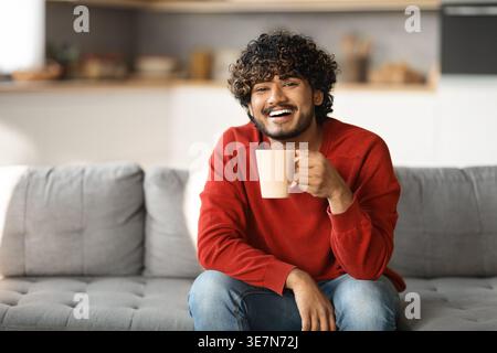 Portrait of happy young indian man with cup of coffee resting on couch at home, young eastern guy sitting on sofa in living room interior, enjoying ho Stock Photo