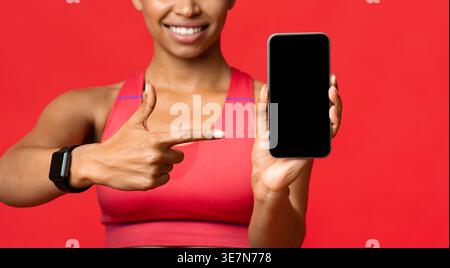 Cropped image of Black woman pointing to smartphone and standing against red background in studio Stock Photo