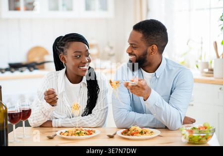 Black couple looking at each other eating pasta sitting at table in kitchen at home Stock Photo