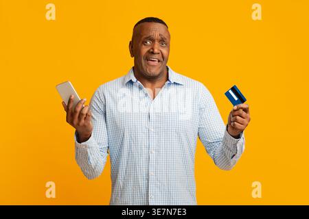 Upset middle-aged Black man holding smartphone and credit card checking his balance while standing against yellow background in studio Stock Photo