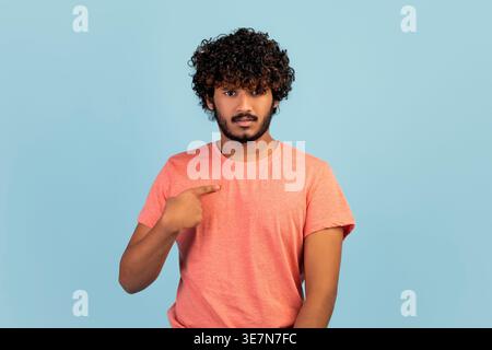 Confused bearded Indian man with curly hair looking away pointing finger at himself while standing in front of blue background in studio Stock Photo