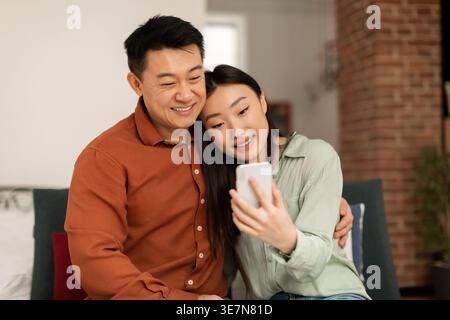 Smiling Asian man hugging wife using smartphone together while sitting on sofa in living room at home Stock Photo