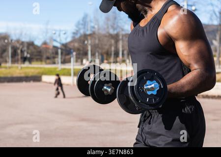 Muscular mixed-race man wearing black tank top and cap lifting dumbbells while standing in park on sunny day Stock Photo