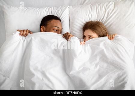 Black and caucasian couple looking at each other hiding on white blanket while lying on bed at home Stock Photo