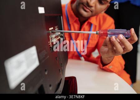 Indian male technician fixing TV using screwdriver while crouching at table in house of customer Stock Photo