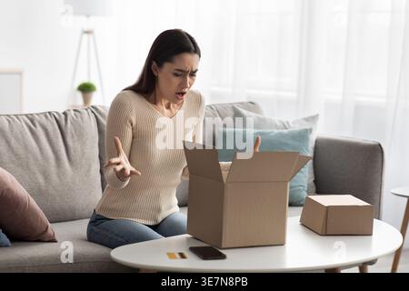Angry Arab woman gesturing hands looking at cardboard box having problem with her package sitting on sofa near table in living room at home Stock Photo