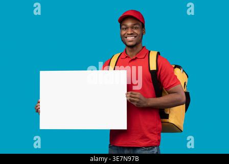 Black delivery man wearing cap carrying thermo backpack holding white blank board standing against blue background in studio Stock Photo