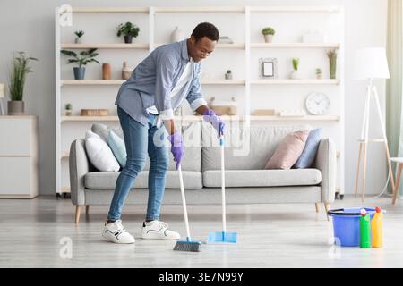Cheerful handsome black guy cleaning cozy living room alone, holding broom and scoop, copy space. Smiling african american young man enjoying house cl Stock Photo