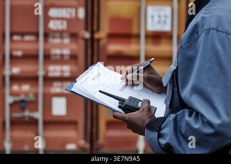 Black male worker holding clipboard and handheld radio standing and checking containers in shipping port Stock Photo