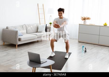 Smiling Asian man wearing white shirt looking at laptop doing deep squats in living room at home Stock Photo