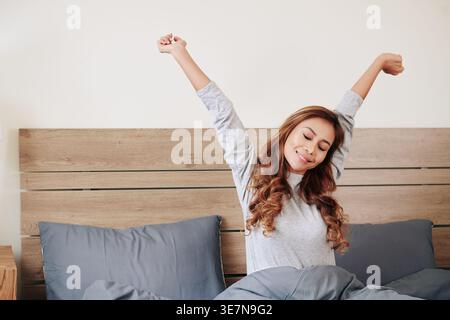 Beautiful happy young Asian woman stretching in bed after waking up in the morning Stock Photo